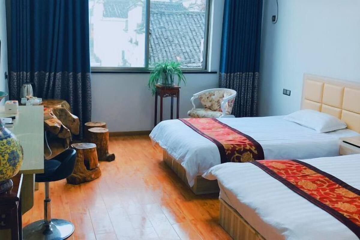 Une chambre d’hôtel avec deux lits, une chaise, une petite table, une plante et une fenêtre avec des rideaux.