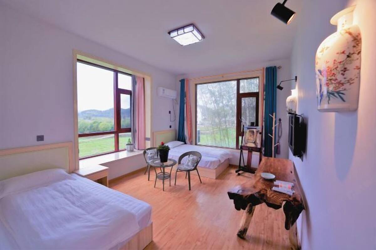 Une chambre avec deux lits, une fenêtre donnant sur un paysage verdoyant, une petite table avec une chaise et une tête d’animal décorative accrochée au mur.