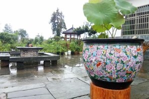 Ein Blumentopf mit floralem Dekor auf einem hölzernen Sockel in einem Garten mit einem Steinfbrunnen und einem Pagodentempel im Hintergrund.