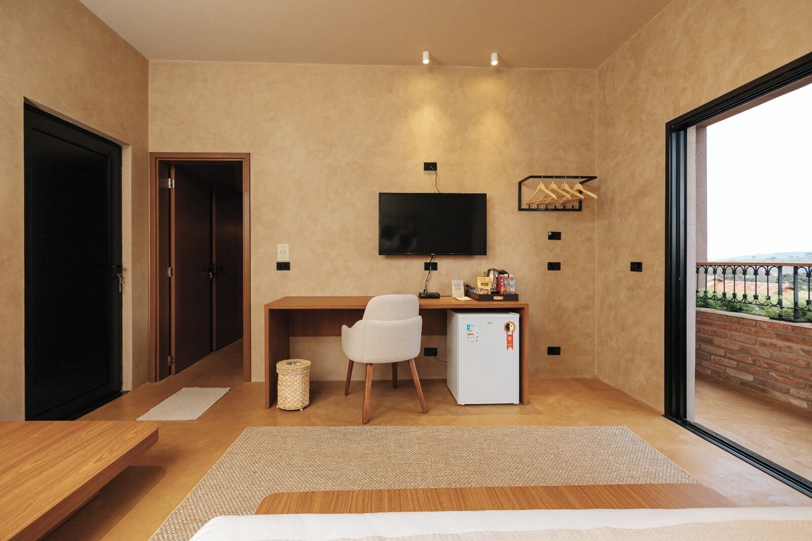 Quarto de hotel moderno com escrivaninha de madeira, uma cadeira branca, TV de tela plana, pequena geladeira e varanda com vista.