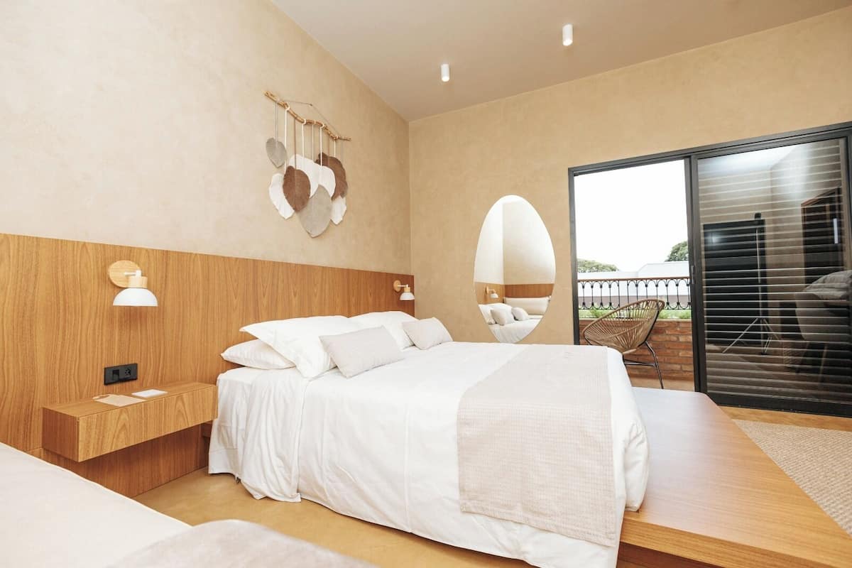 Ein modernes Schlafzimmer mit Holz-Kopfteil, einem Nachttisch, einem großen Bett mit weißer Bettwäsche, einer Wandleuchte und Blick ins Freie durch eine Schiebetür.