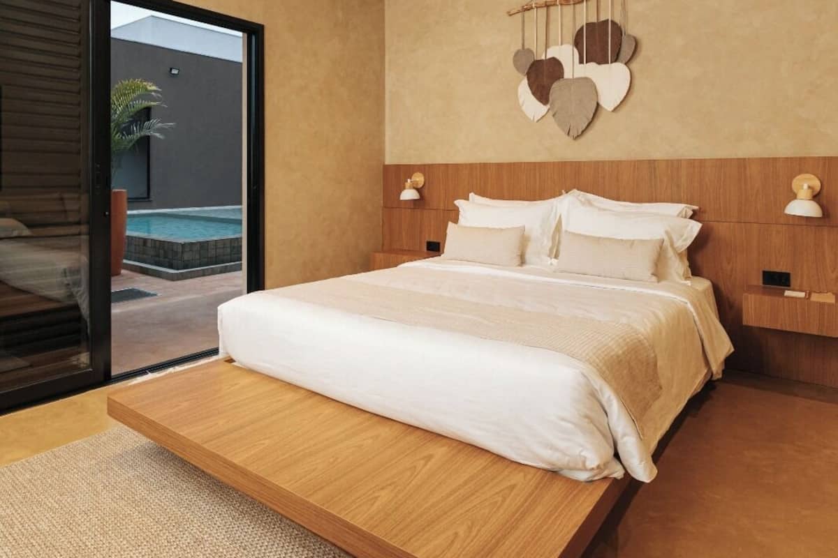 Ein modernes Schlafzimmer mit einem großen Bett, einem Holz-Kopfteil und Blick auf einen Swimmingpool durch eine Glastür.