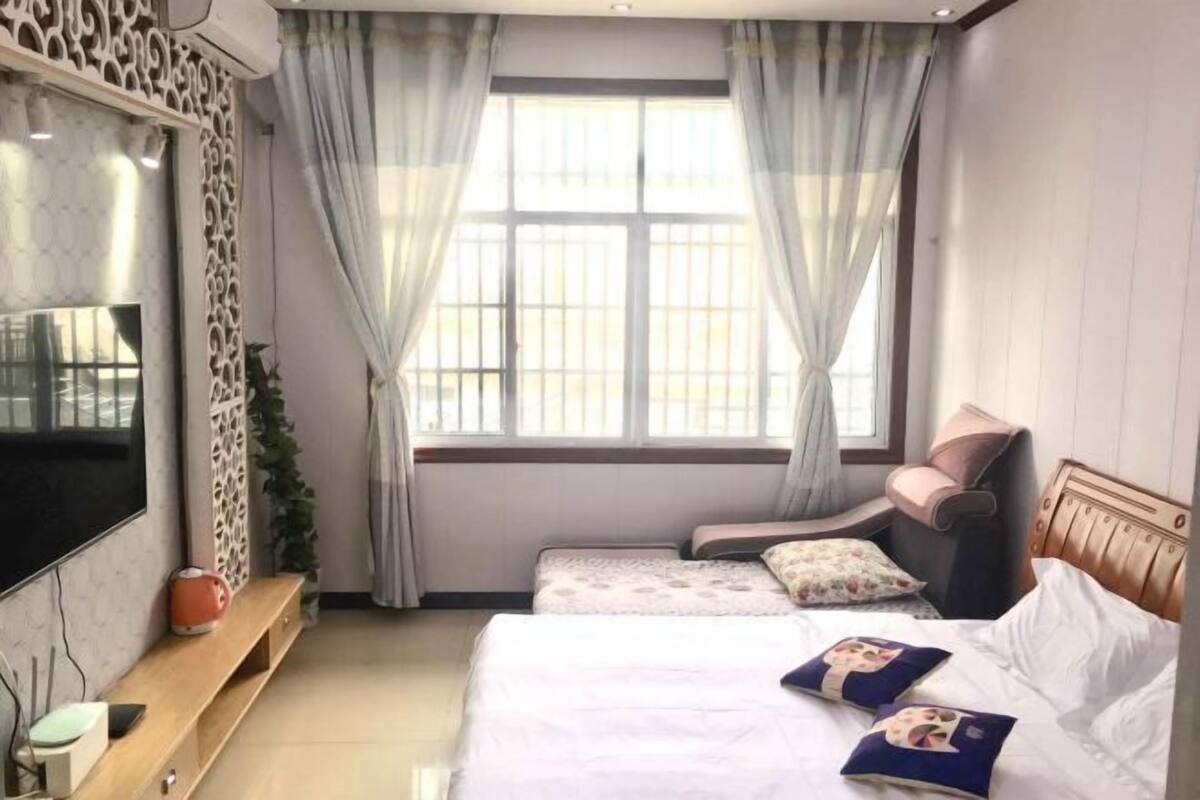 A hotel room with a large flat-screen TV, two beds with decorative pillows, a wooden bench, and a window with curtains.