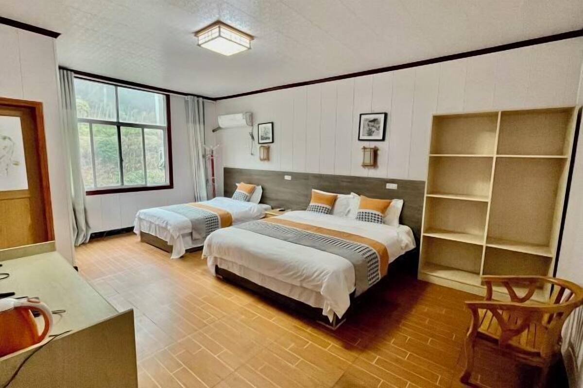 A hotel room with two beds, a wooden floor, a large window, and a bookshelf.