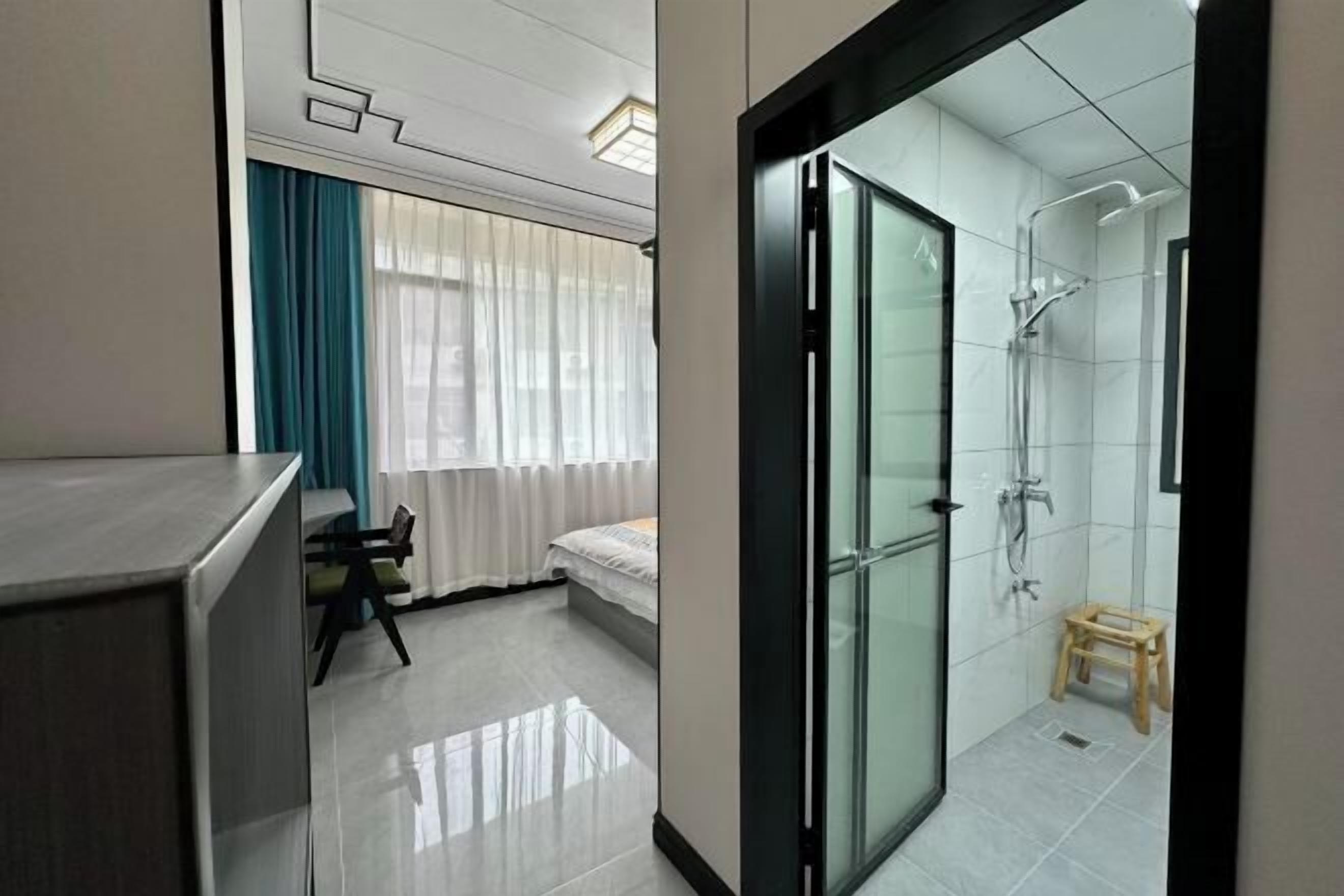 A modern hotel room with a glass shower enclosure, a bed, a small wooden stool, and a dining area with a table and chairs.