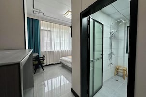 Habitación de hotel moderna con cabina de ducha de vidrio, cama, taburete de madera pequeño y zona de comedor con mesa y sillas.