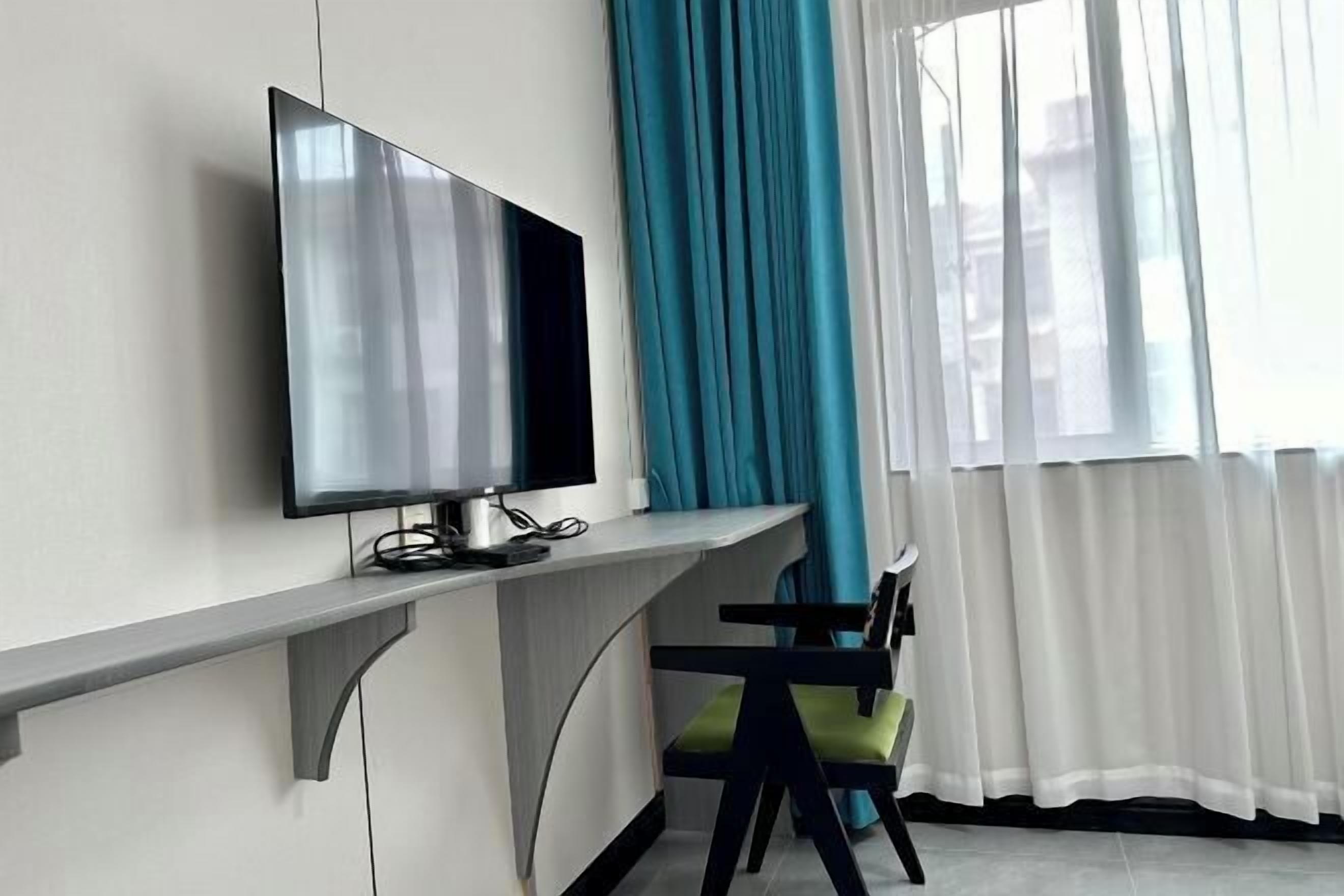 A modern room with a flat-screen TV mounted on a wall, a desk with a chair, and a window with sheer white curtains.