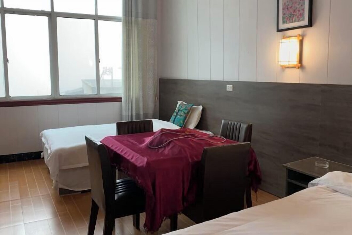 A hotel room with two beds, a dining table with chairs, a small bedside table, and a window with curtains.