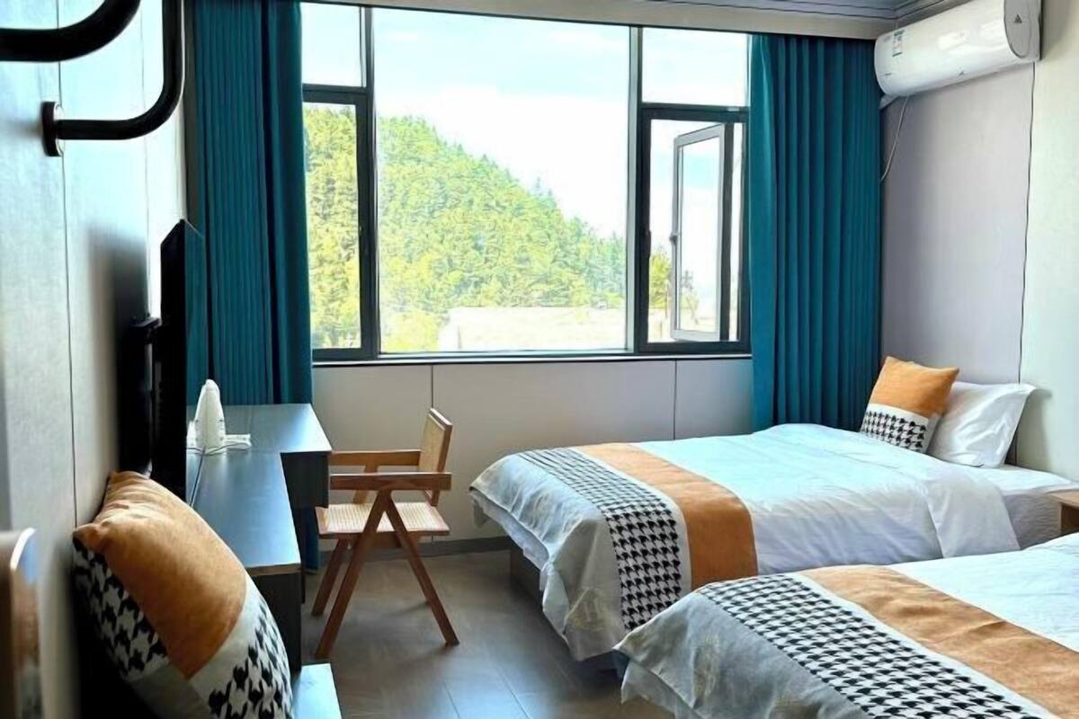 A hotel room with two beds, a desk, and a chair. There is a large window with a view of a forested area.