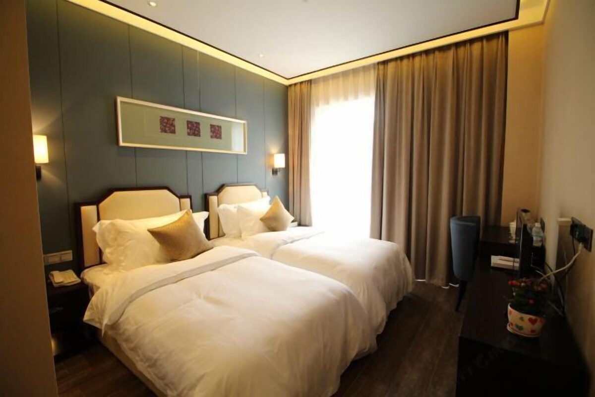 Quarto de hotel com duas camas, uma mesa de trabalho, uma cadeira, uma luminária e uma janela com cortinas.