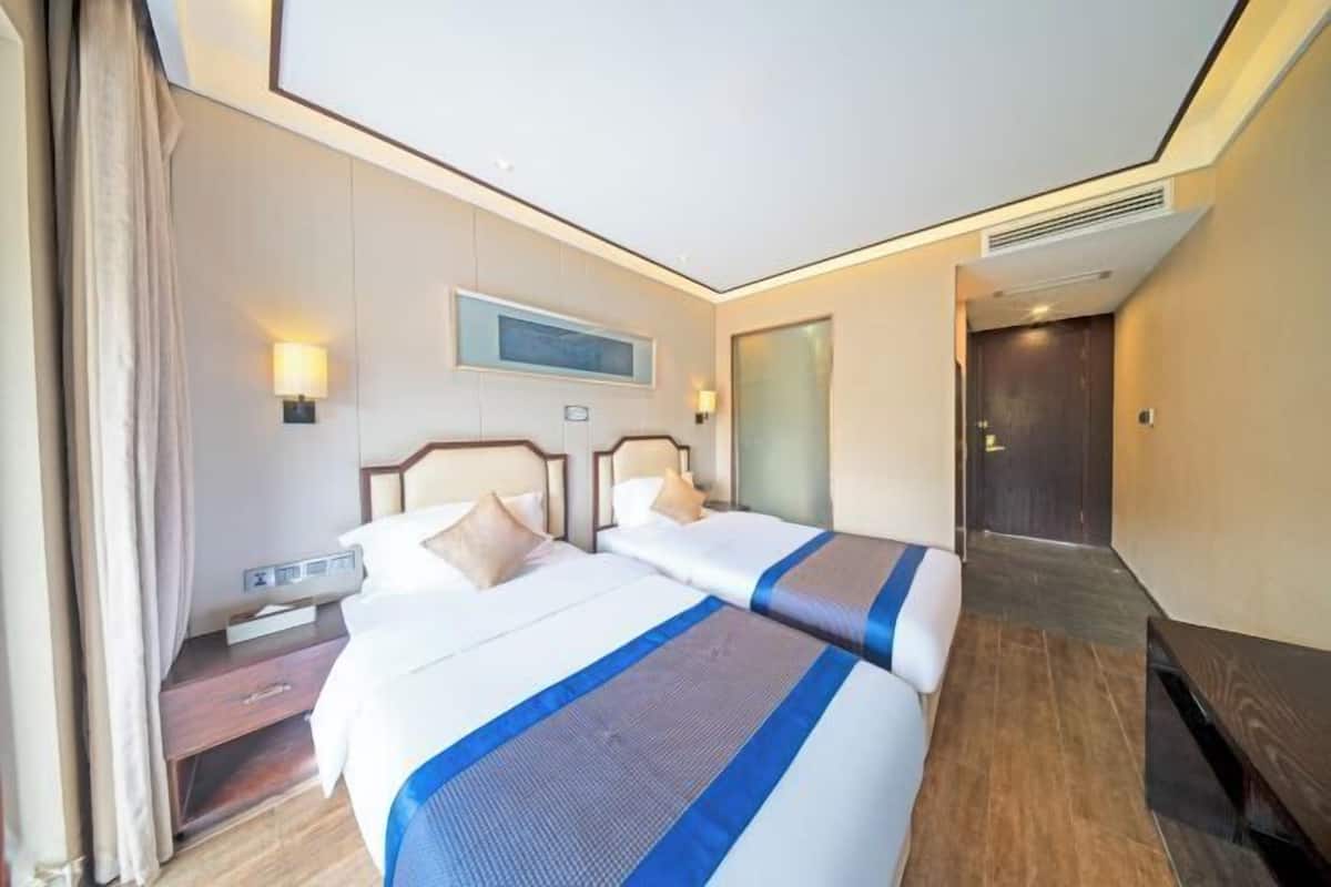 Quarto de hotel com duas camas, mesa de cabeceira, escrivaninha, cadeira e porta.
