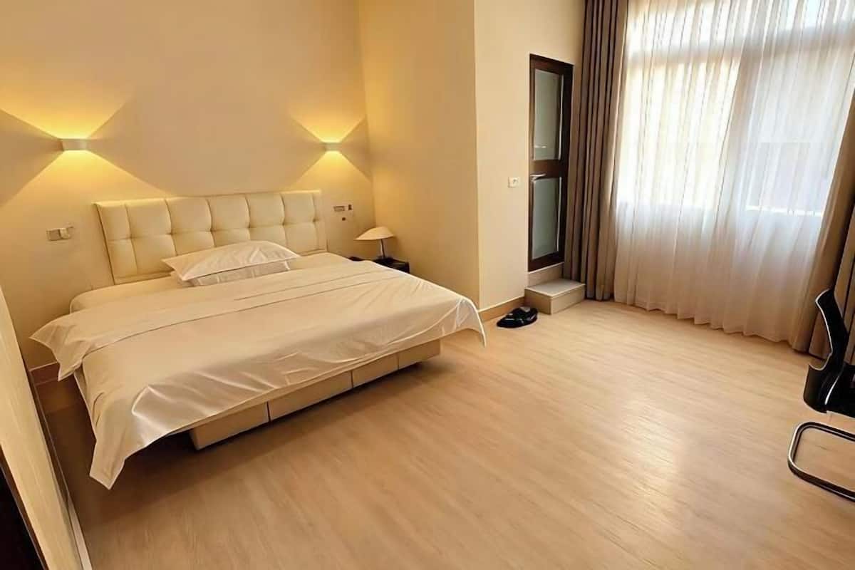 Una habitación de hotel con cama, lámparas de noche, una silla, ventana con cortinas y una puerta.