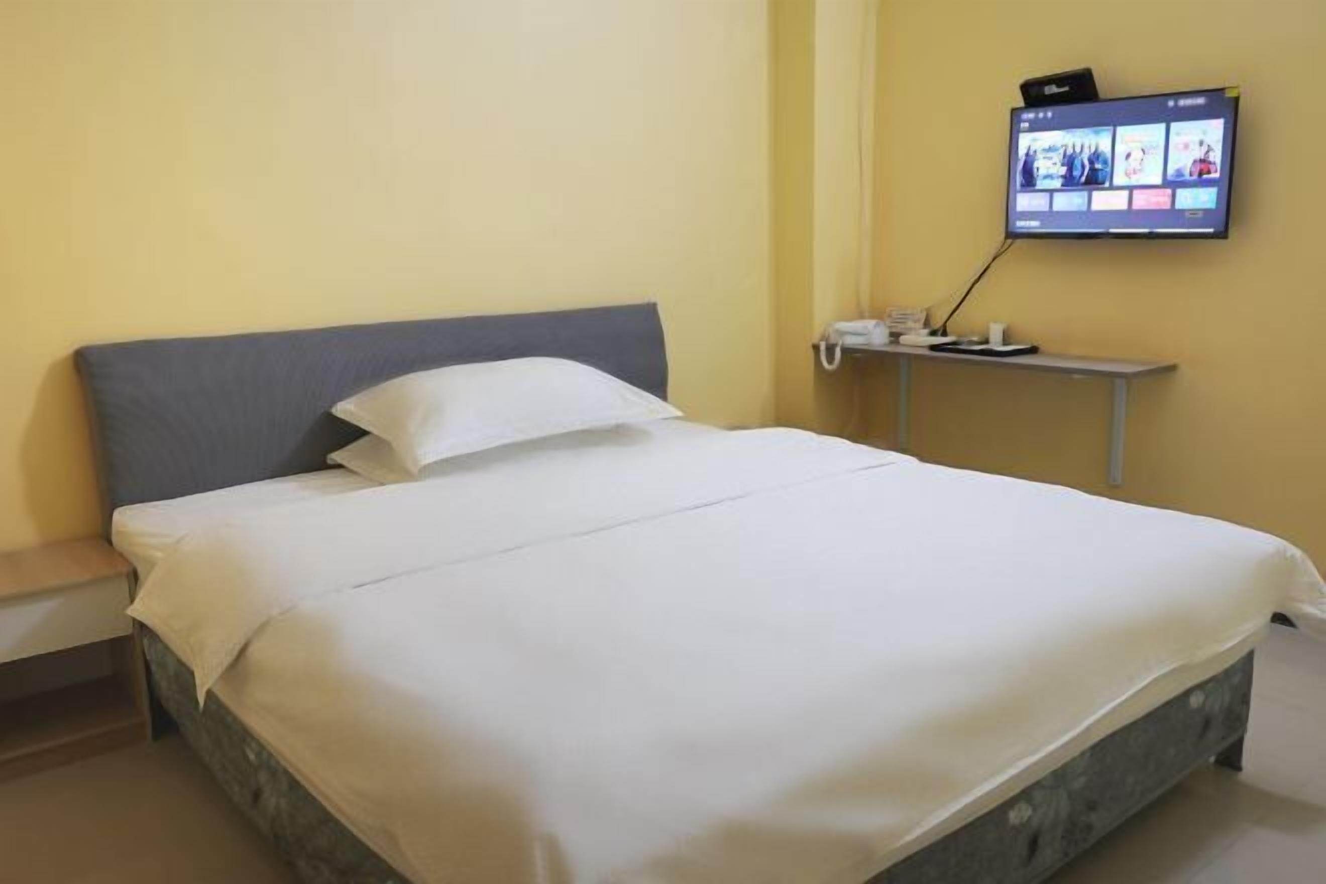 A neatly made bed with white linens in a room with a yellow wall and a flat-screen TV mounted on the wall.