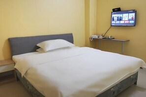 Un lit bien fait, avec du linge de lit blanc, dans une pièce dont un mur est jaune et où un téléviseur à écran plat est fixé au mur.