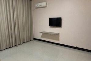 Una habitación con cortina, aire acondicionado, televisor de pantalla plana montado en la pared y un estante pequeño.