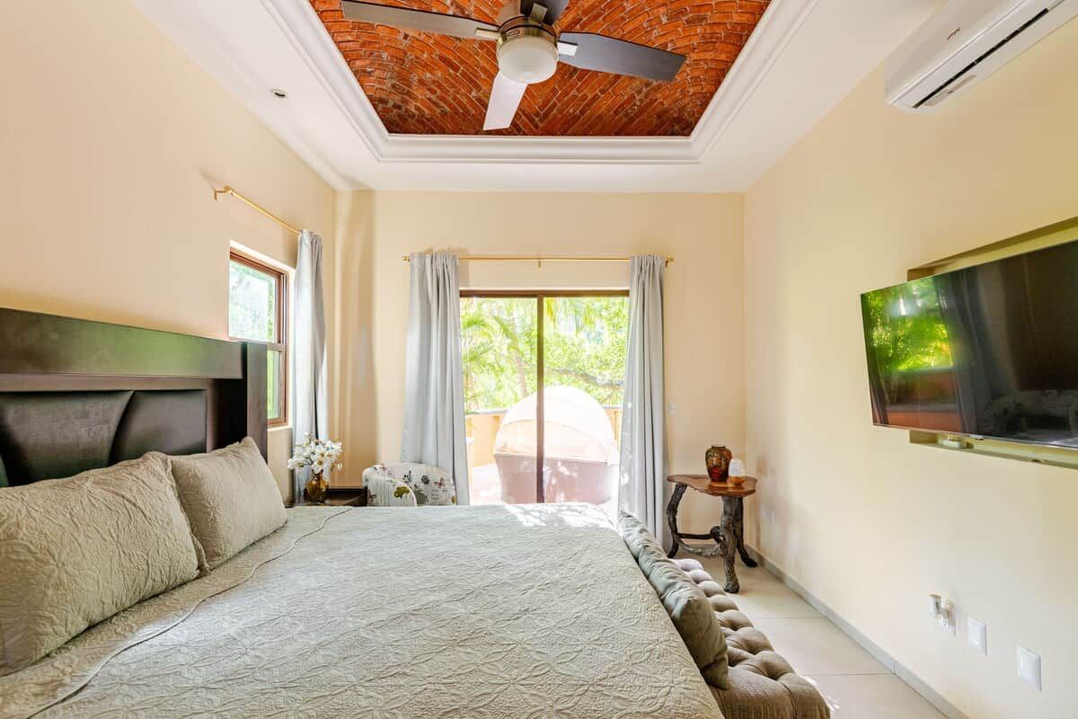 A bedroom with a bed, a ceiling fan, a TV mounted on the wall, and a window with curtains.