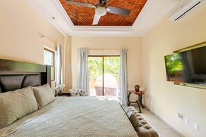 Un dormitorio con una cama, ventilador de techo, televisor montado en la pared y una ventana con cortinas.