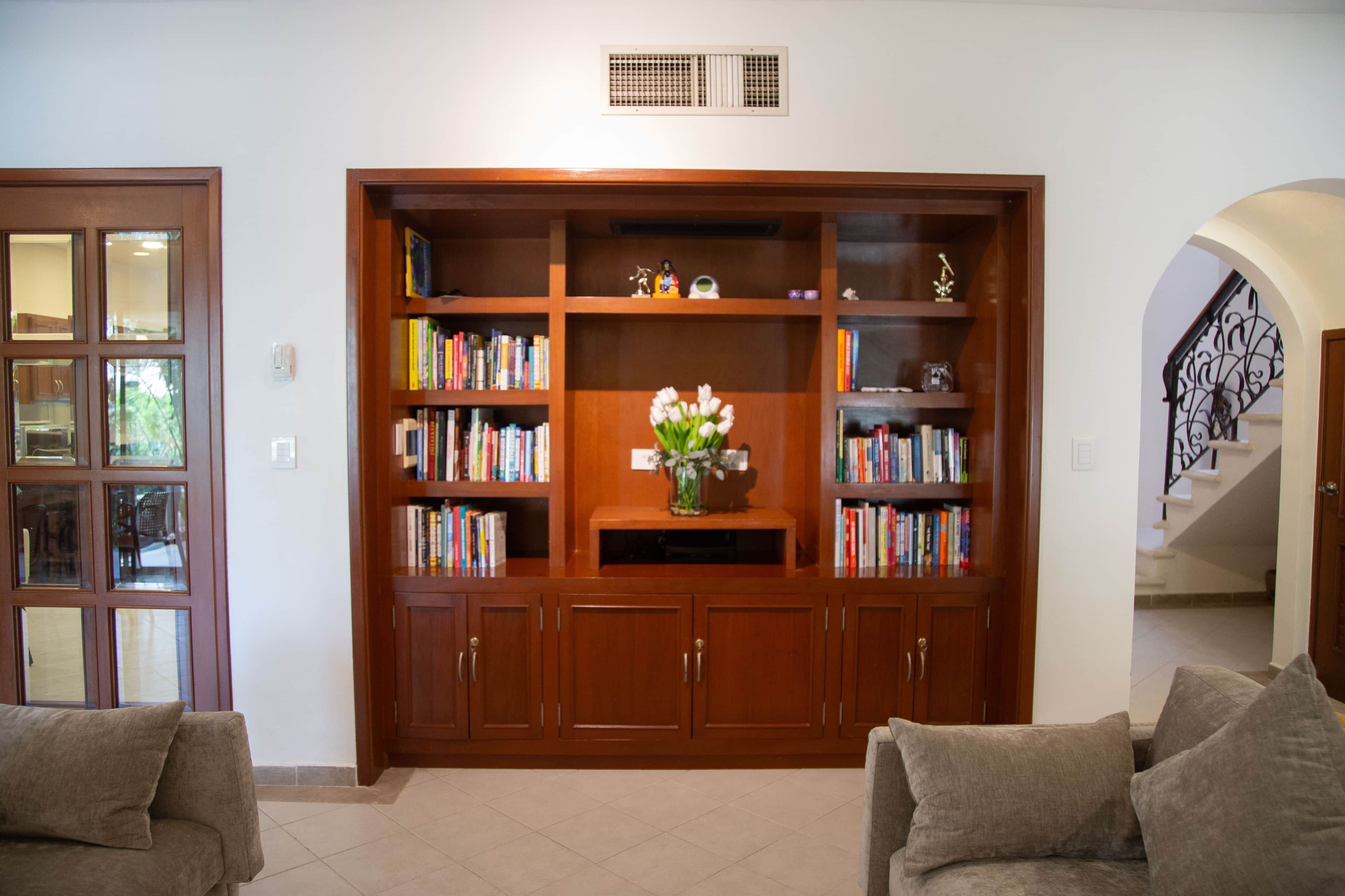 A living room with a wooden bookshelf, a vase with flowers, and a staircase in the background.