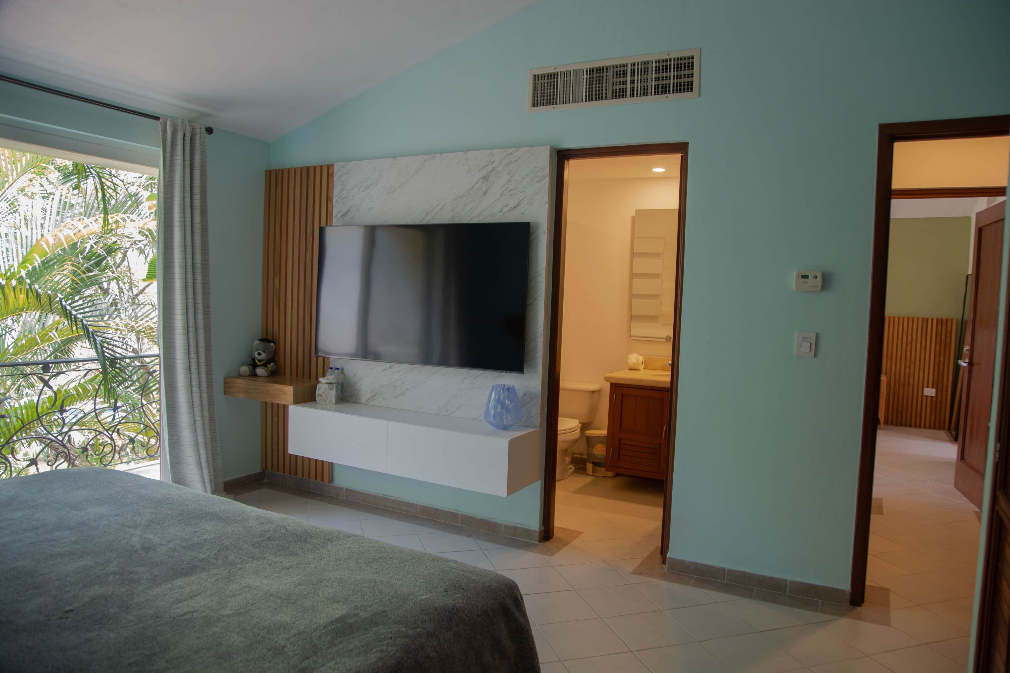 Quarto de hotel moderno com uma televisão de tela plana grande fixada numa parede de mármore, uma cama com cobertura cinza, uma pequena prateleira com um urso de pelúcia e vista para o exterior através de uma janela ampla.