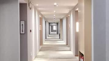 Corredor longo e estreito de um hotel, com piso coberto por carpete e um extintor de incêndio na parede direita.