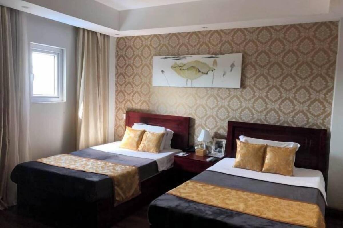 A hotel room with two beds, a window with curtains, a patterned wallpaper, and a painting on the wall.
