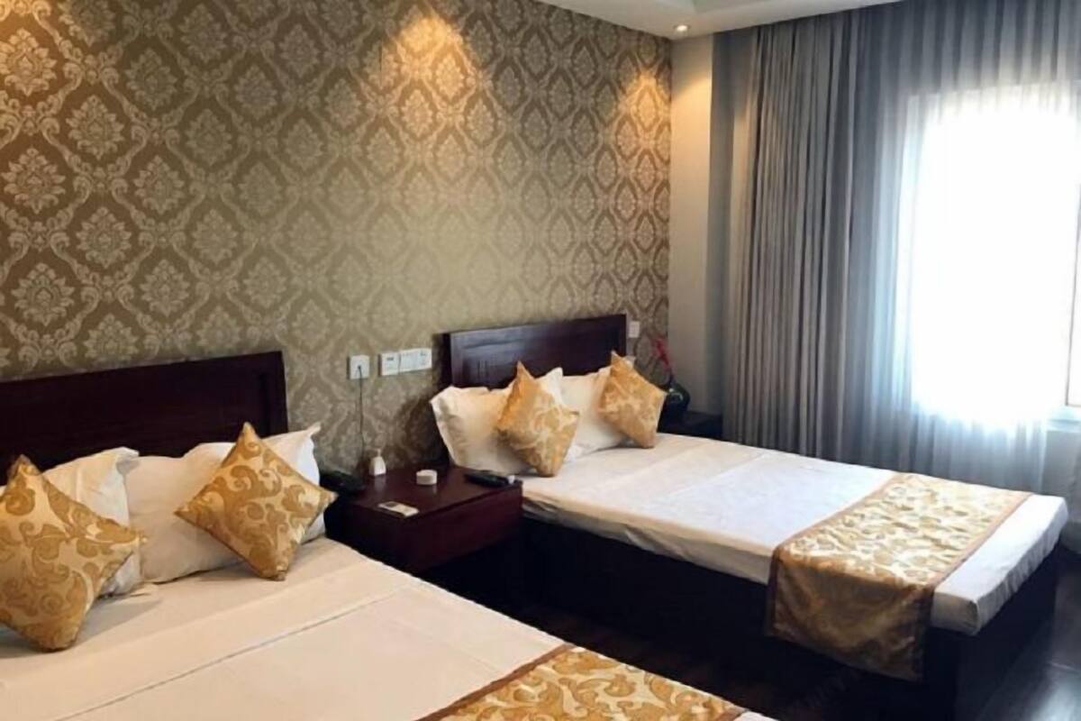 A hotel room with two beds, patterned wallpaper, a dark wooden headboard, and a window with curtains.