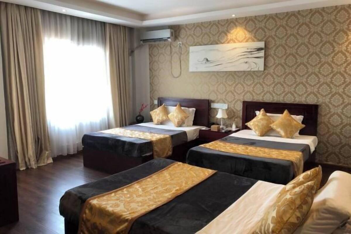 A hotel room with two beds, a large window with curtains, a wall-mounted air conditioner, and a painting.