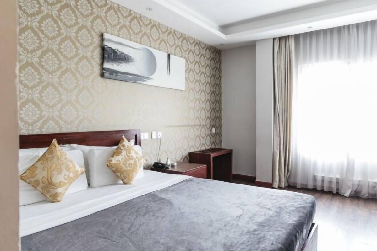 A hotel room with a bed, a nightstand, a wall-mounted artwork, and a window with curtains.