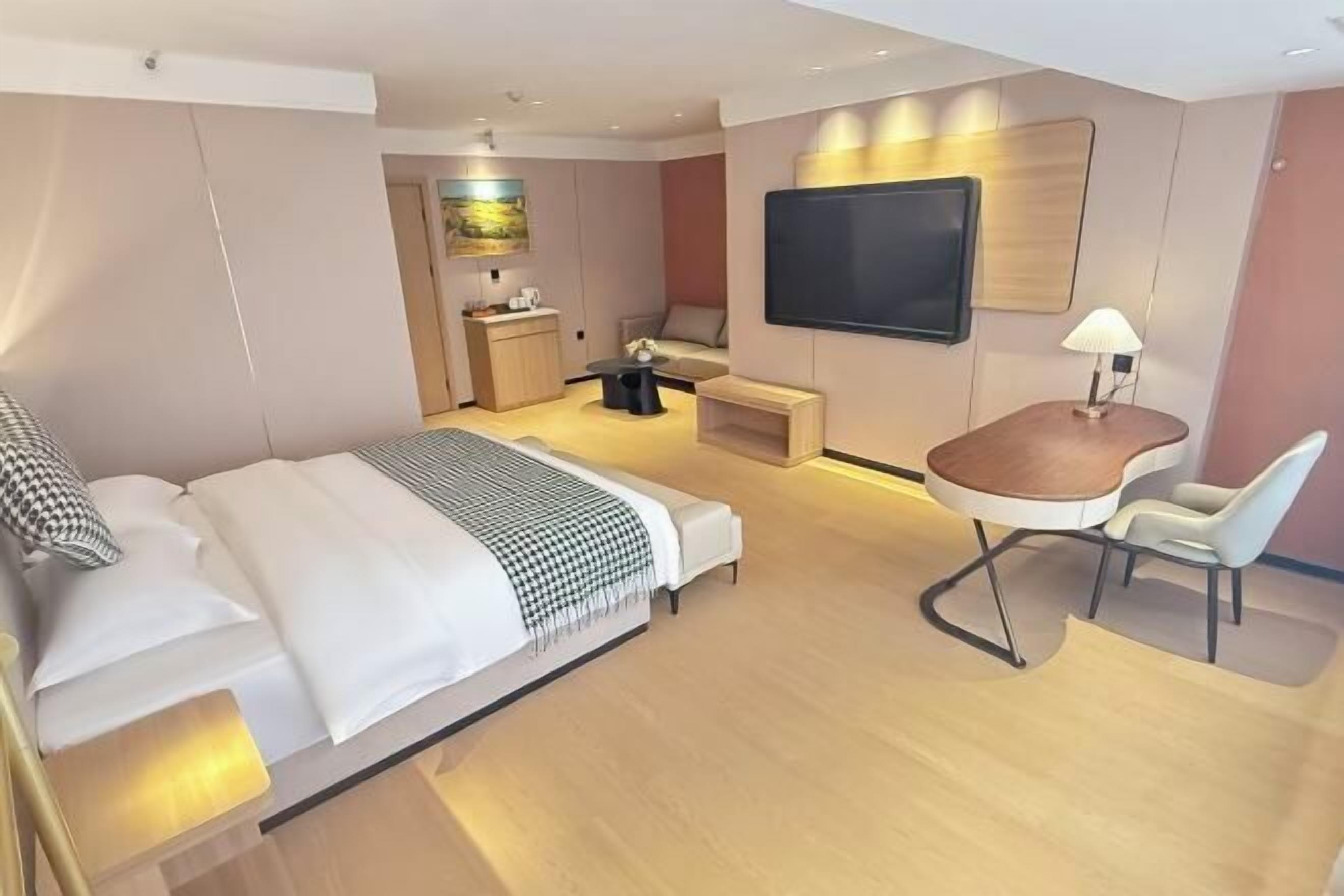 A modern hotel room with a bed, a desk, a chair, a TV, and a small table with a lamp.
