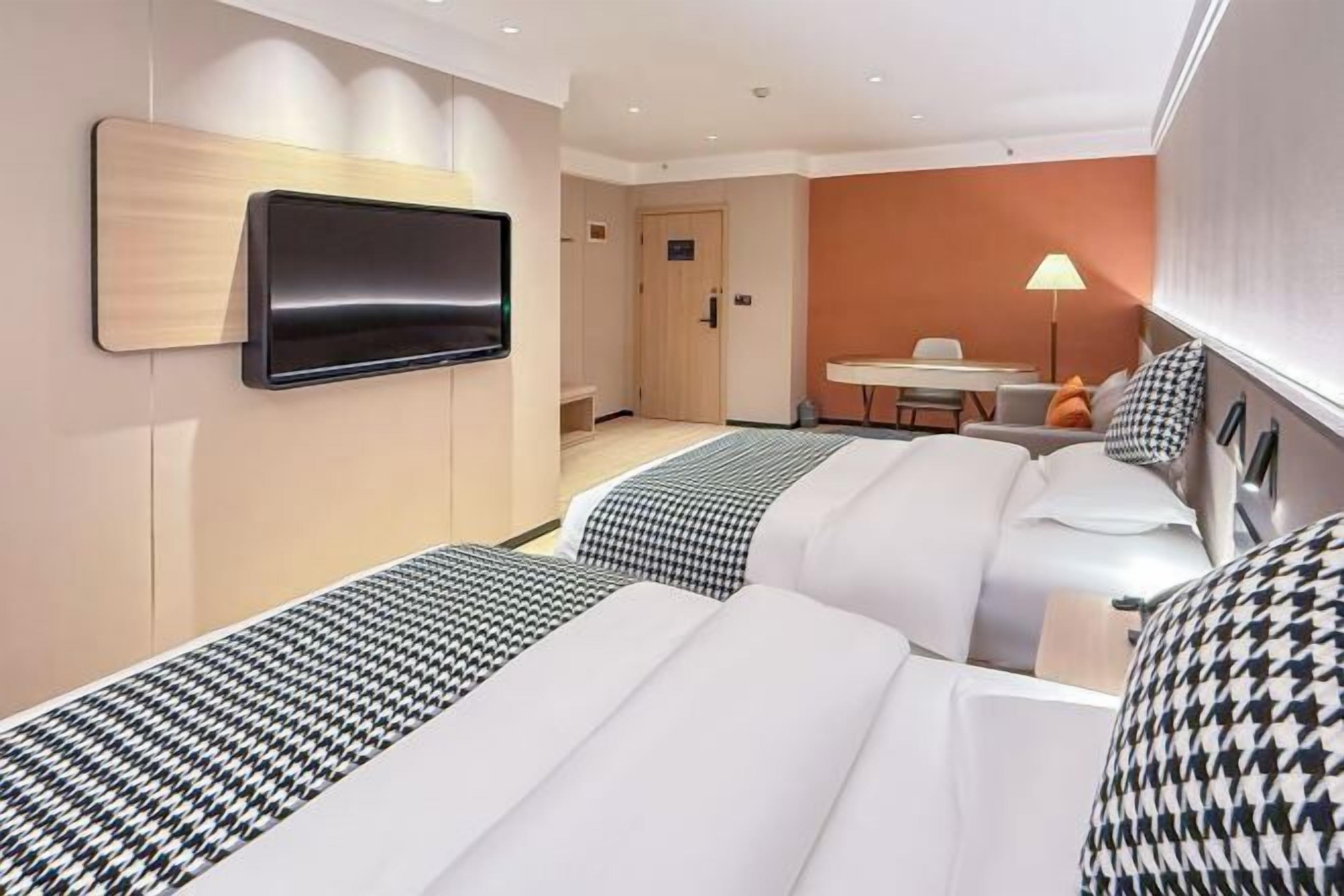 A hotel room with two beds, a TV mounted on the wall, a desk with a chair, and a lamp.