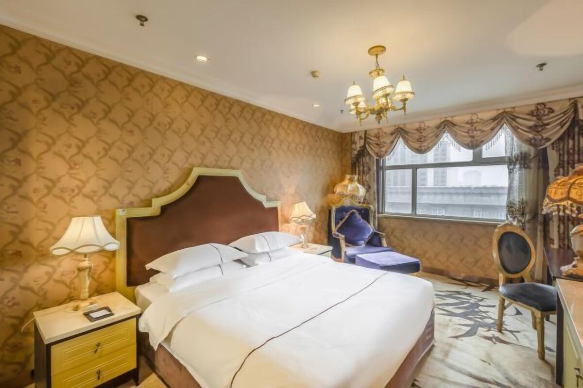 A hotel room with a large bed, a nightstand, a lamp, a chair, and a window with curtains.