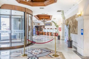 Un vestíbulo de hotel con entrada de vidrio, mostrador de recepción, candelabro y alfombra estampada.
