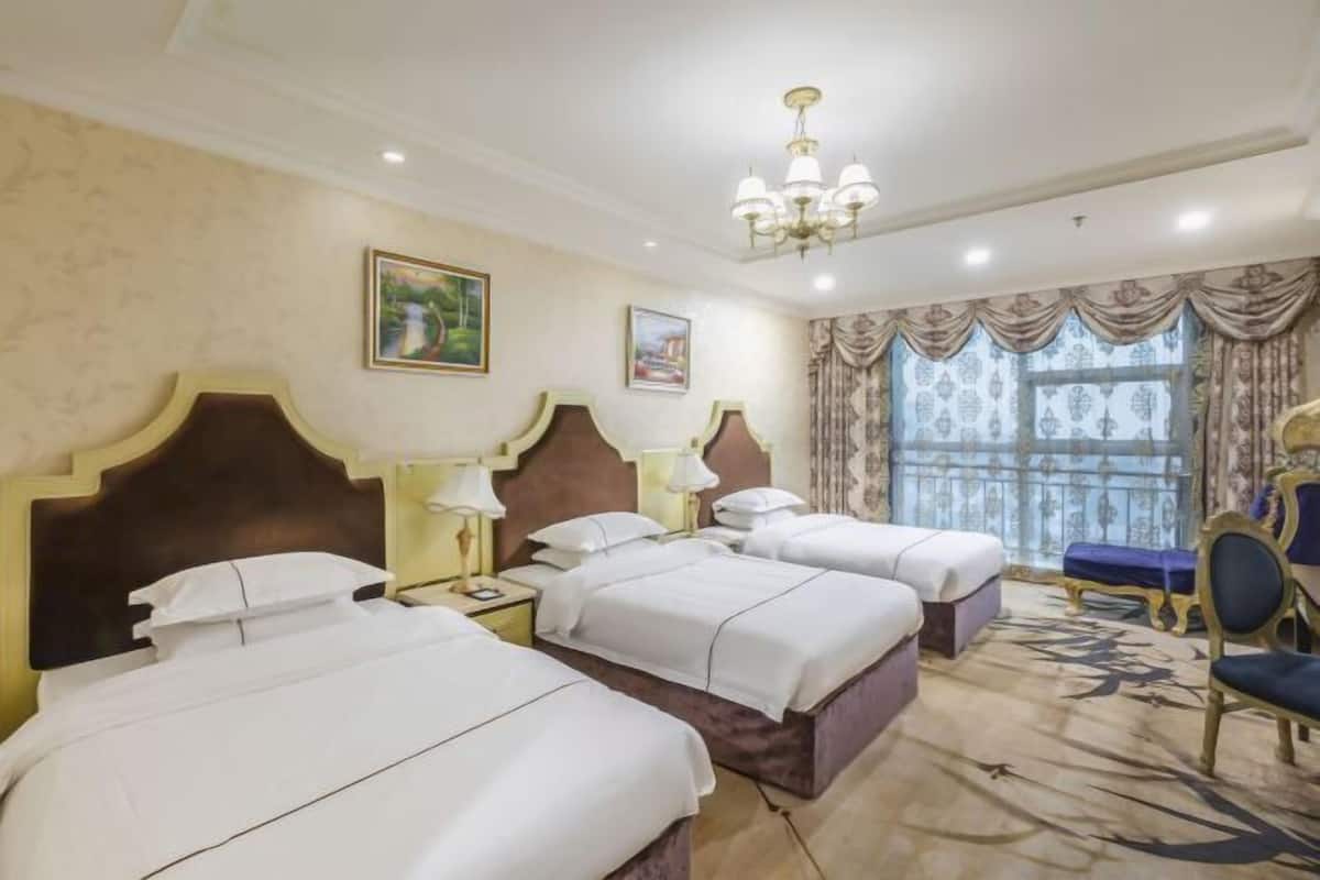 Habitación de hotel con tres camas, un candelabro y un gran ventanal con cortinas.