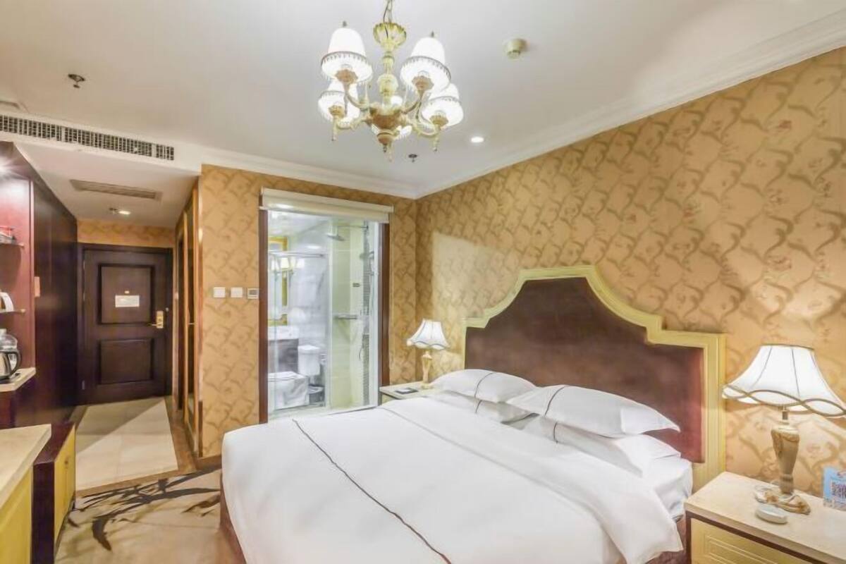 Habitación de hotel con una cama grande, un candelabro, una mesita de noche y un baño visible a través de una puerta abierta.