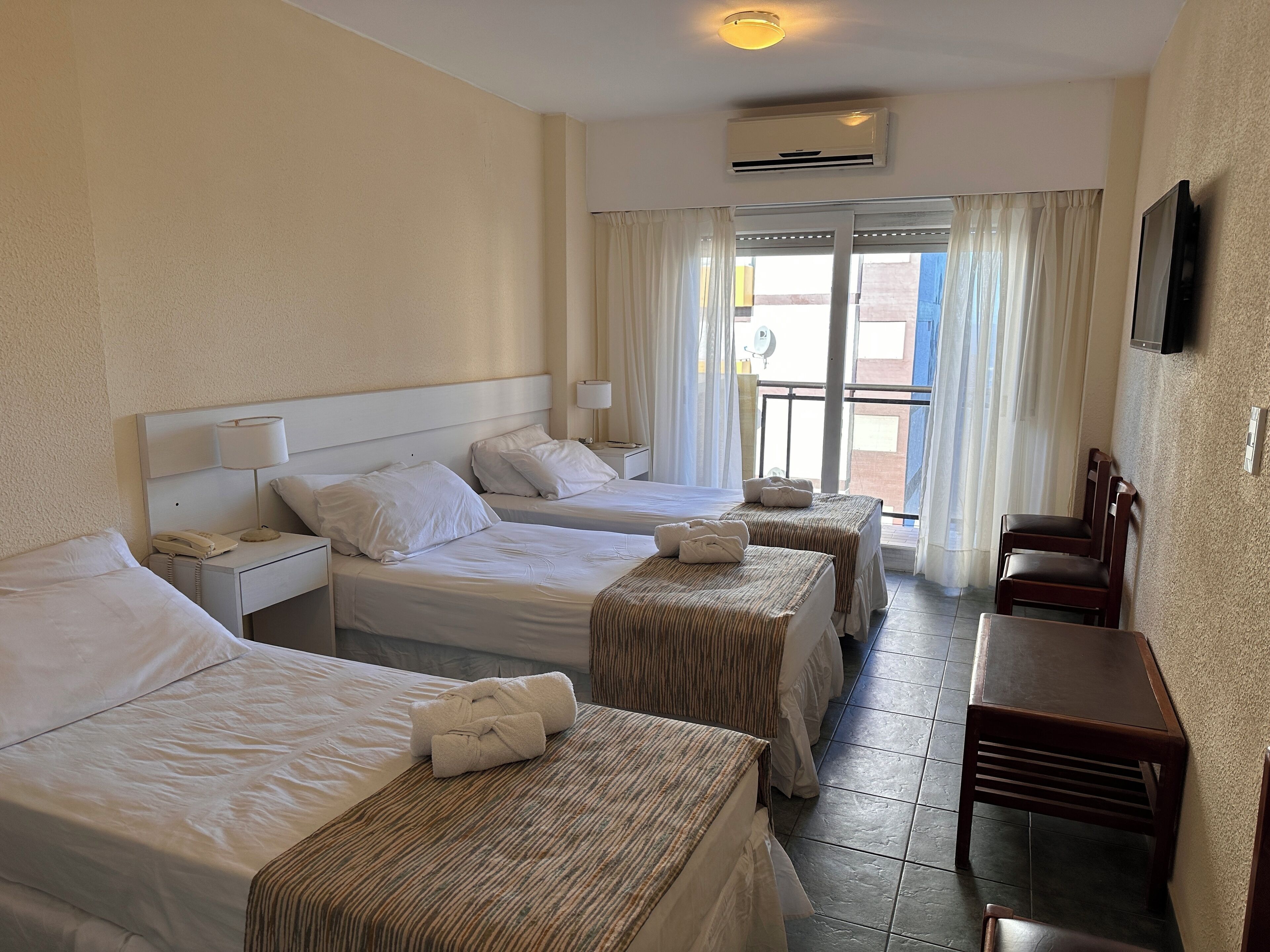 Ein Hotelzimmer mit zwei Betten, einem Schreibtisch und einem Stuhl. An der Wand befindet sich eine Klimaanlage und ein Fenster mit Vorhängen.