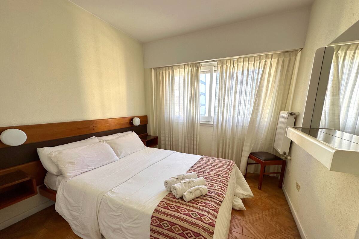 A hotel room with a wooden headboard bed, white bedding, a patterned throw, a small wooden stool, and a window with sheer curtains.