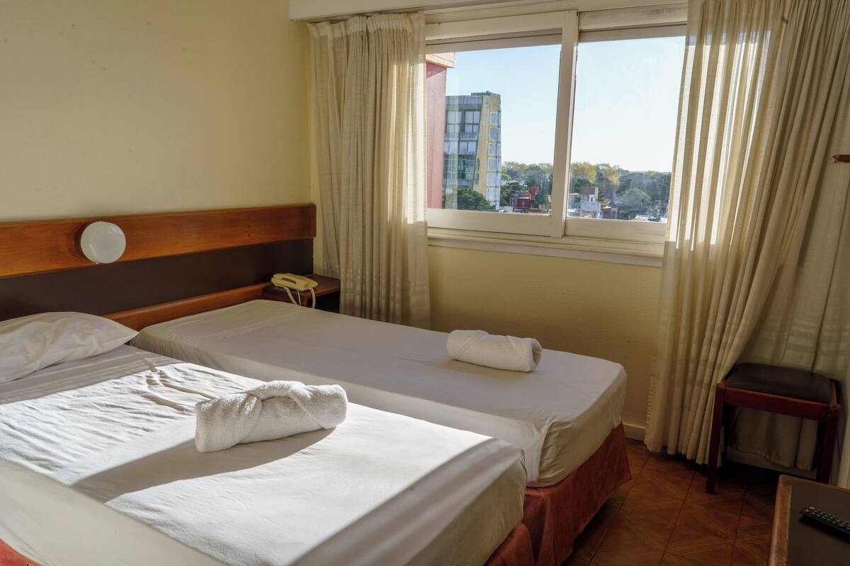 A hotel room with two beds, a wooden headboard, a nightstand, a window with curtains, and a view of buildings outside.