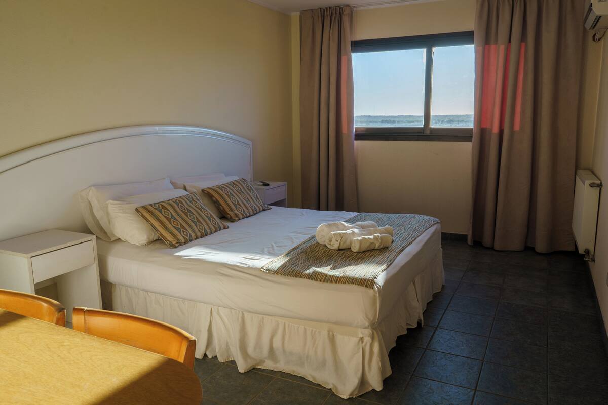 A neatly made bed with pillows, a bedside table, a chair, and a window with curtains.