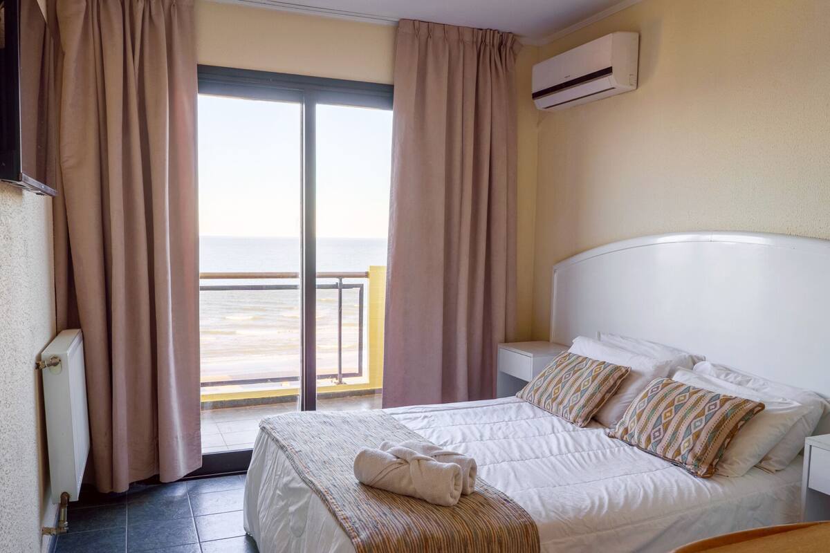 A hotel room with a bed, a view of the sea through a sliding door, and an air conditioning unit on the wall.