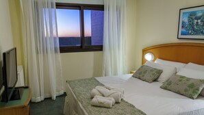 A hotel room with a neatly made bed, a window with curtains, a bedside table with a lamp, and a painting on the wall.