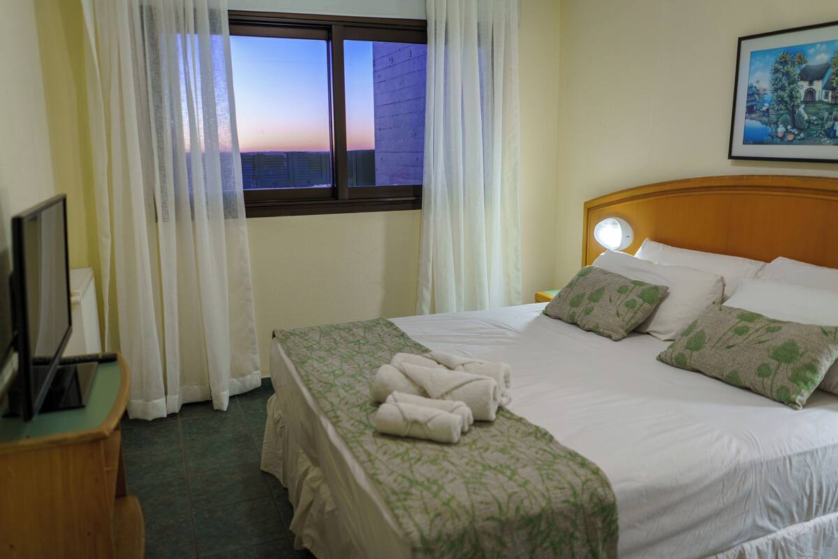 A hotel room with a neatly made bed, a window with curtains, a bedside table with a lamp, and a painting on the wall.