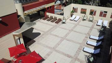 Terraza en la azotea con sillones de sol y sombrillas rojas, un área de estar cubierta y vistas de edificios circundantes.