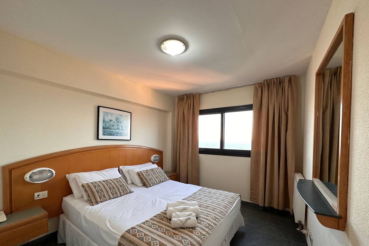 A hotel room with a bed, a nightstand, a bedside table, a window with curtains, a ceiling light, and a framed picture on the wall.