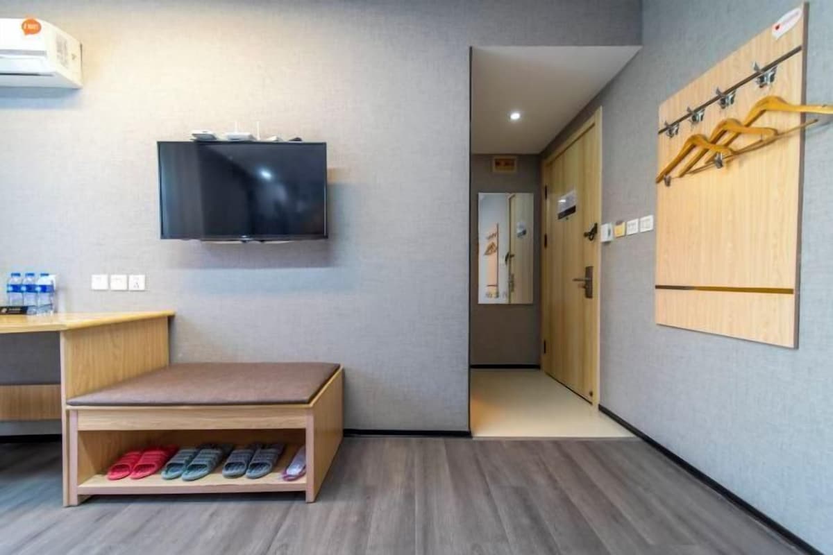 A hotel room with a TV mounted on the wall, a wooden bench with slippers, a shoe rack, and a wooden wardrobe.