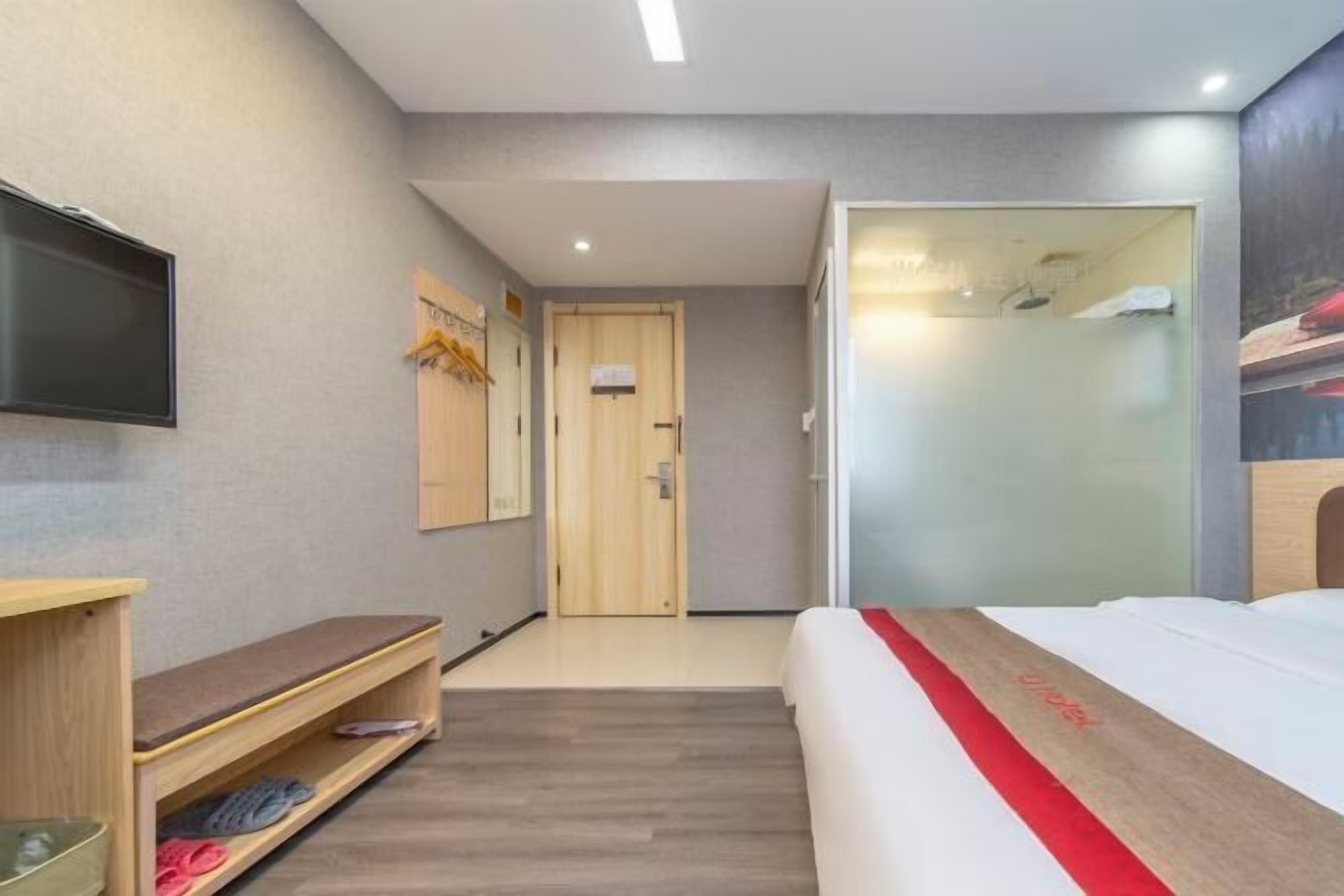 A hotel room with a bed, a TV mounted on the wall, a wooden bench with slippers, and a door leading to a bathroom.
