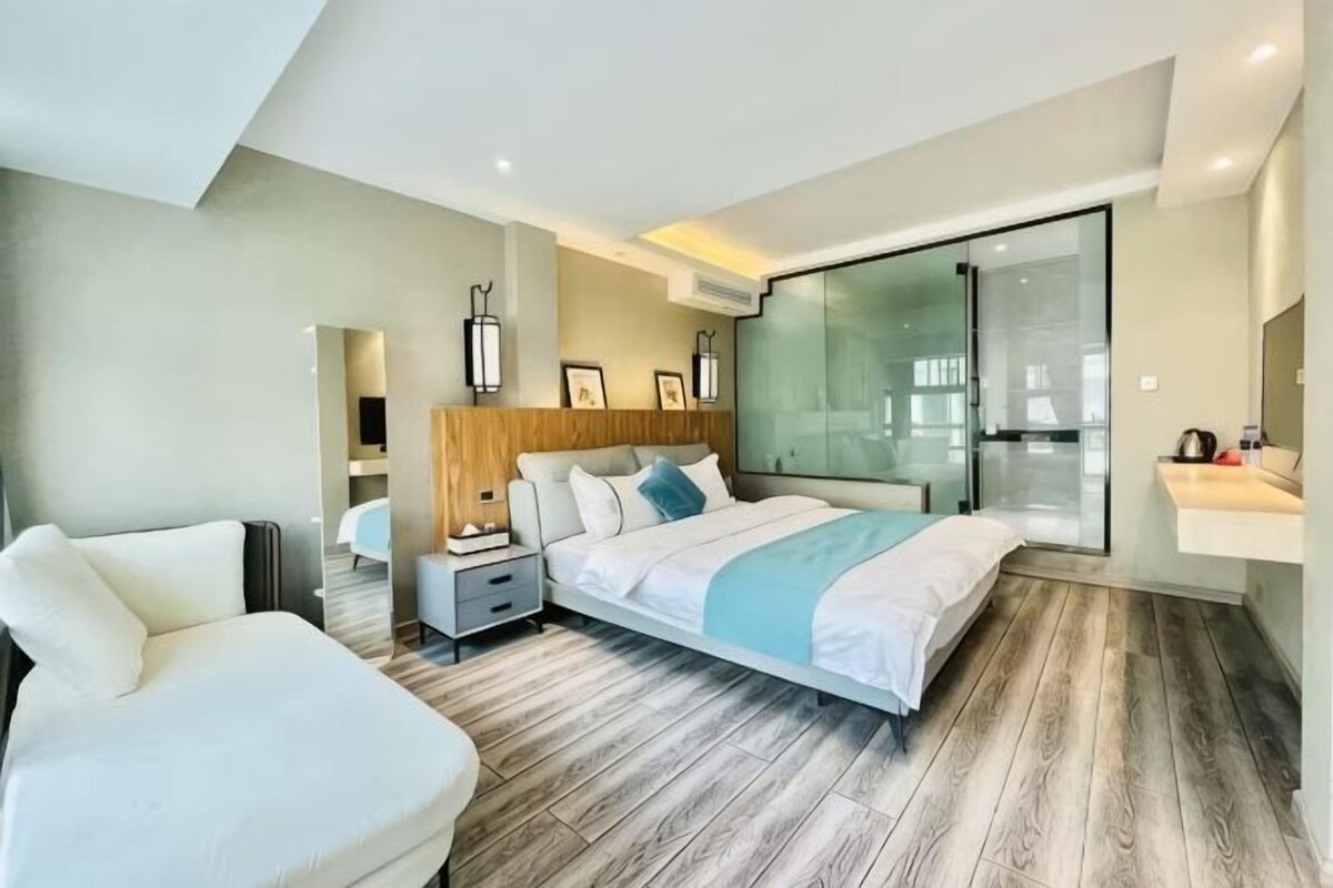 A modern hotel room with a large bed, a seating area, a glass door leading to a balcony, and a small desk with a lamp.