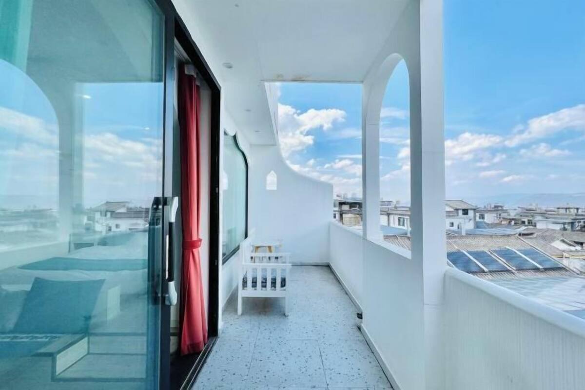 A narrow balcony with a glass door, a small white bench, and a view of a residential area with houses and solar panels.