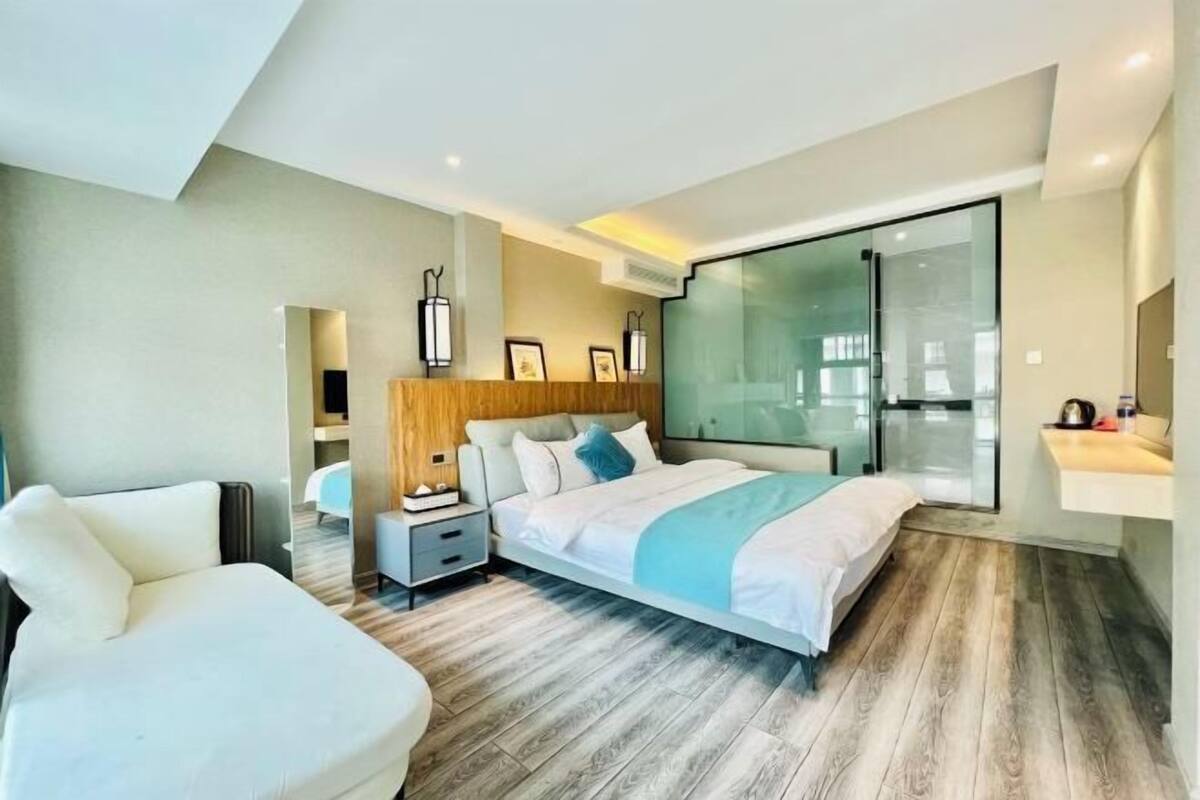 A modern hotel room with a large bed, a seating area, a glass partition, and a small table with a kettle.