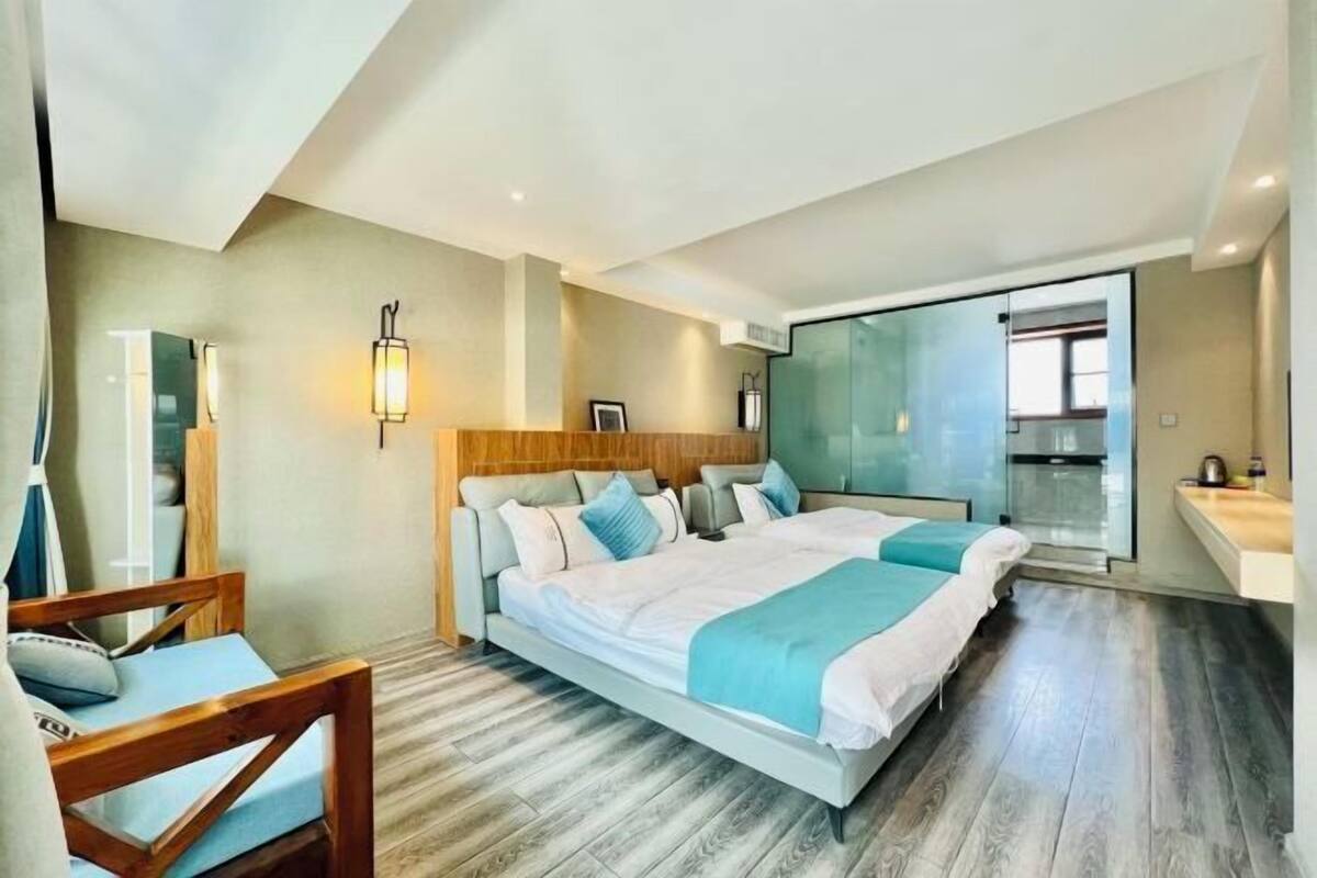 A modern hotel room with a large bed, a wooden headboard, a glass shower enclosure, and a small kitchenette.