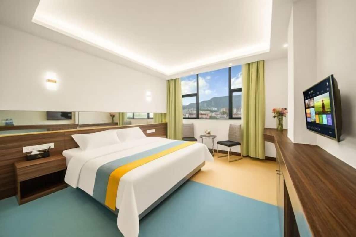 A hotel room with a bed, bedside table, a small table with chairs, a TV, and a large window with a city view.